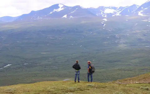 Två vandrare njuter av utsikten över vidsträckta fjäll och skogar, kanske under en vandra abisko 3 dagar. Snöklädda berg tornar upp sig i bakgrunden.