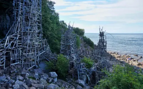 Nimis i Skåne: en skulptur av trä som klättrar uppför klipporna vid havet.