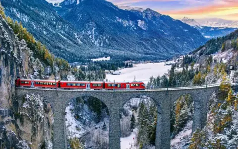 Röda tåget korsar en stenbro över en snöig dal i Schweiz. Fantastiska vyer för din resa till Schweiz tips.