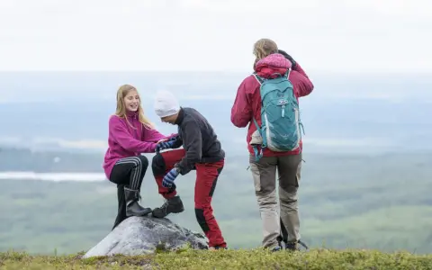 Två barn hjälper varandra upp på en sten under en vandring. En vuxen står bakom med ryggsäcken redo för äventyr.