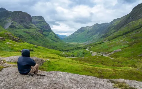 Person sitter på en sten och njuter av utsikten över en grön dal i Skottland. En perfekt plats för att vandra i Skottland.