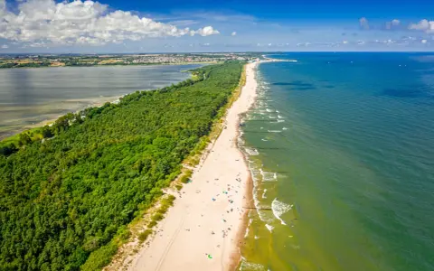 En lång sandstrand kantad av en grönskande skog. Havet glittrar i solljuset och en stad syns i bakgrunden. En perfekt sommardag på hel polen.