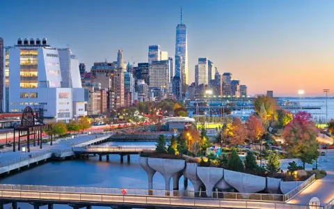 Fakta om New York: En vy över Manhattans skyline vid solnedgången, med den ikoniska One World Trade Center och en modern park vid vattnet.
