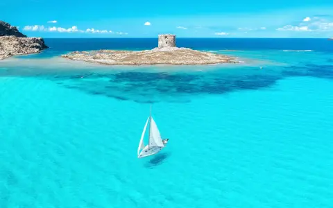En segelbåt glider fram på turkost vatten mot en gammal vakttorn på en ö. Perfekt för din resa till Sardinien tips!
