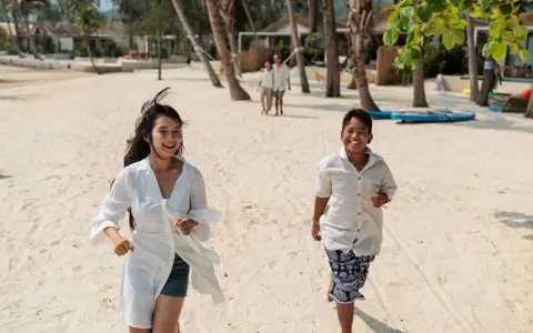 Två barn springer glatt på en sandstrand i Thailand. I bakgrunden syns palmer och bungalows.