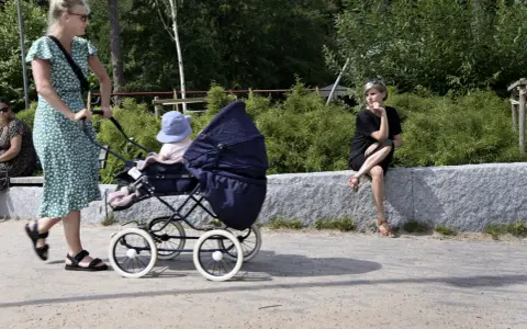 En kvinna med barnvagn njuter av sin semester i Sverige med barn. En annan kvinna sitter på en stenmur i bakgrunden.