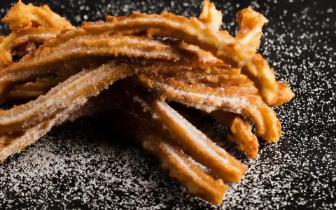 En hög med gyllene, sockerbeströdda churros, en härlig friterad deg, ligger på en mörk yta.