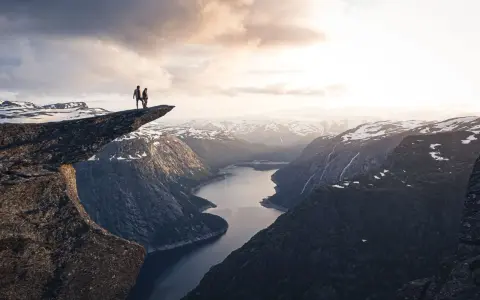 Två personer står på Trolltunga efter en 3 dagars vandring i Norge. Dramatisk utsikt över fjord och snöklädda berg.
