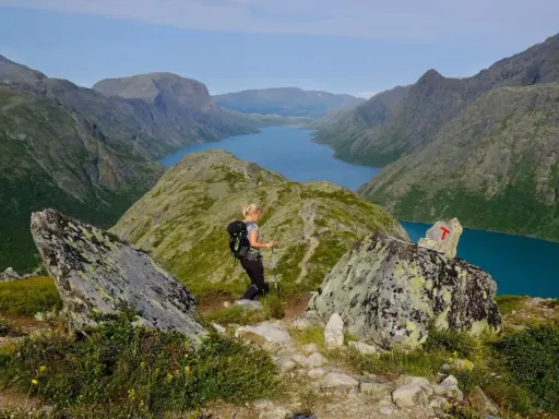 En kvinna vandrar i Jotunheimen med en blå sjö och berg i bakgrunden. En T-märkt sten visar vägen.
