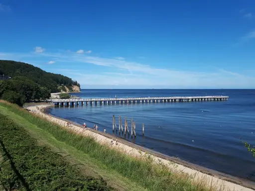 En lång pir sträcker sig ut i det blå havet vid Gdynia, Polen. En grön kulle med träd reser sig vid sidan av stranden där några människor promenerar.