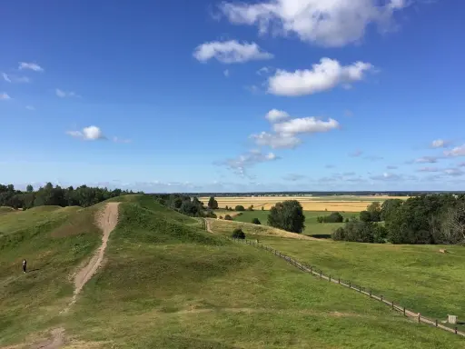 Gröna kullar med stigar och en ensam person. Ett vackert landskap att se i Uppsala med fält och skog i bakgrunden under en blå himmel med moln.