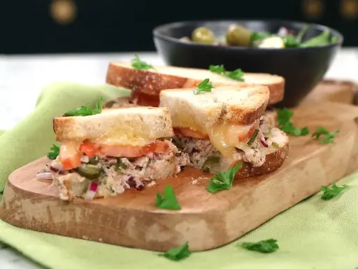 Två halvor av en gyllene tuna melt med smält ost, tomat och kapris, garnerad med persilja. Serveras på en träskärbräda med en sallad i bakgrunden.