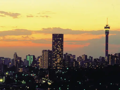 Stadssilhuett i Johannesburg, Sydafrika, med upplysta skyskrapor och ett högt torn mot en gul och orange solnedgångshimmel.