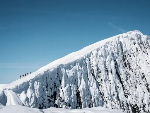 En grupp vandrare på en snöig bergstopp under en helags topptur. Den isiga bergväggen reser sig dramatiskt mot en klarblå himmel.