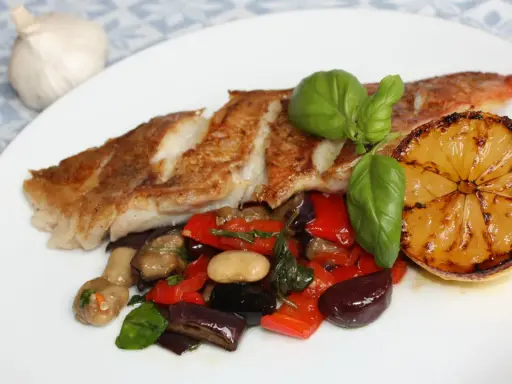 Pan-seared fish with roasted vegetables, a grilled lemon slice, and basil on a white plate.