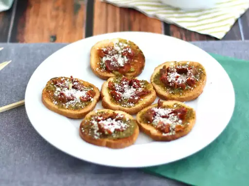 Små, rostade brödskivor toppade med grönt pesto, soltorkade tomater och riven ost. Perfekt som en enkel pesto toast.