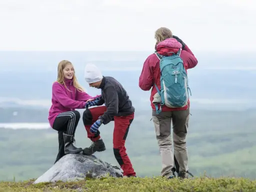 Två barn hjälper varandra upp på en sten under en vandring. En vuxen står bakom med ryggsäcken redo för äventyr.