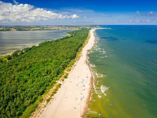En lång sandstrand kantad av en grönskande skog. Havet glittrar i solljuset och en stad syns i bakgrunden. En perfekt sommardag på hel polen.