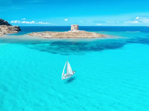 En segelbåt glider fram på turkost vatten mot en gammal vakttorn på en ö. Perfekt för din resa till Sardinien tips!
