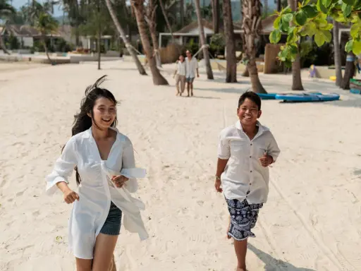 Två barn springer glatt på en sandstrand i Thailand. I bakgrunden syns palmer och bungalows.