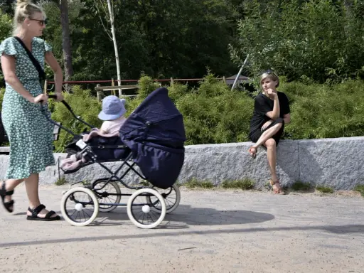 En kvinna med barnvagn njuter av sin semester i Sverige med barn. En annan kvinna sitter på en stenmur i bakgrunden.
