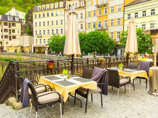 Uteservering vid en flod i Karlovy Vary med gula byggnader i bakgrunden.