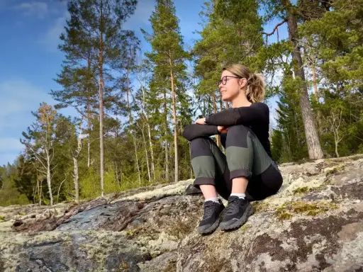 En kvinna vilar på en klippa under en vandring. Solen skiner genom träden, perfekt för vandring rundslingor Stockholm.
