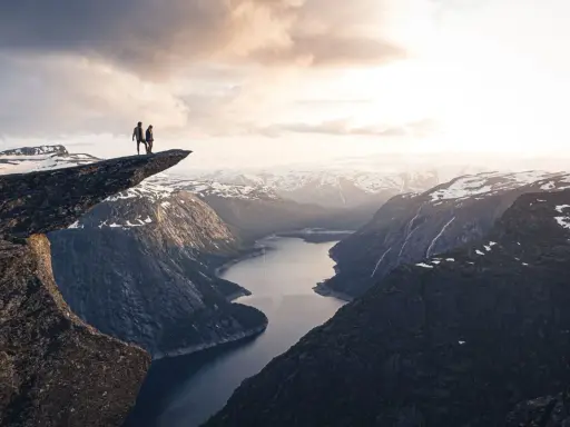 Två personer står på Trolltunga efter en 3 dagars vandring i Norge. Dramatisk utsikt över fjord och snöklädda berg.