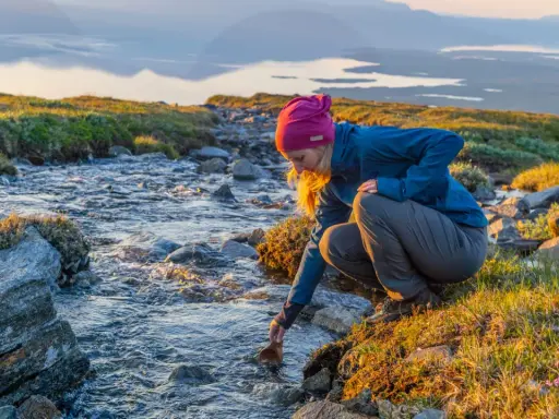 En kvinna fyller sin träkopp vid en bäck under en Abisko vandring dagstur. Solen lyser upp det vidsträckta landskapet.