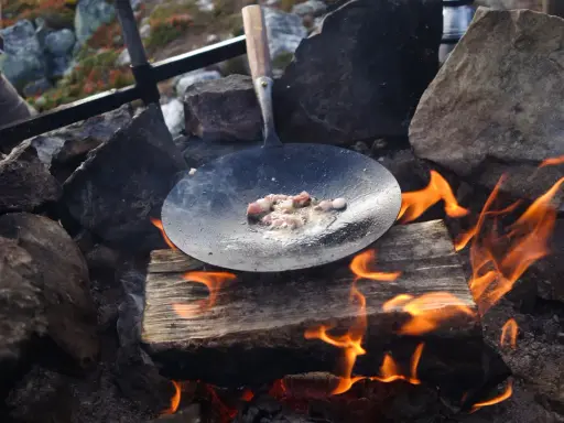 Stekt fläsk i stekpanna över öppen eld, en del av ett recept kolbullar. Elden sprakar och röken stiger.