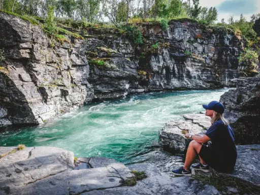 En person sitter vid en forsande turkos flod omgiven av klippor. Ett perfekt utflyktsmål i Mellansverige för naturupplevelser.