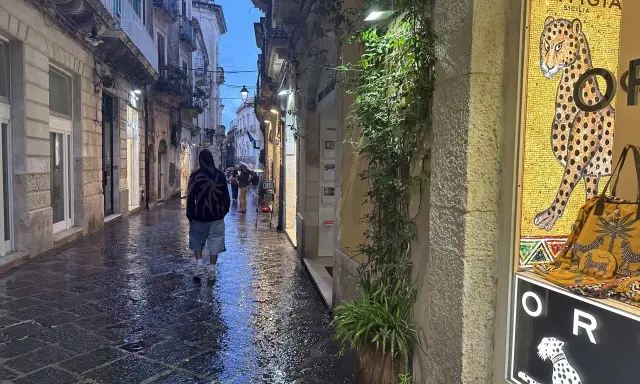 En person går längs en regnvåt gata i Syrakusa, Sicilien. En butiks skyltfönster visar en mosaik av en jaguar och texten "Ortigia".