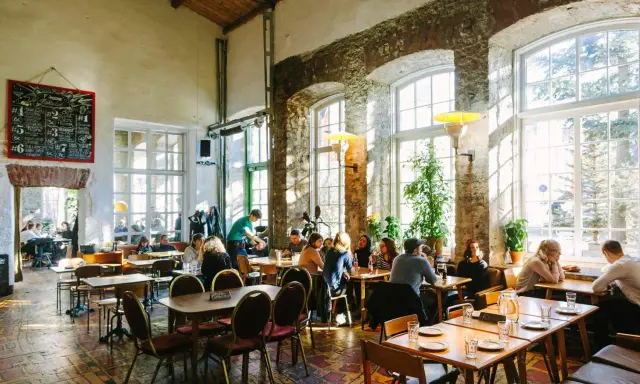 Folk äter och dricker på en livlig restaurang i Tallinn. Stora fönster släpper in solljus i lokalen med rustika tegelväggar.