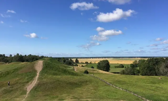 Gröna kullar med stigar och en ensam person. Ett vackert landskap att se i Uppsala med fält och skog i bakgrunden under en blå himmel med moln.