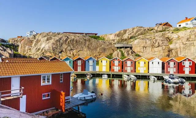 Färgglada sjöbodar längs en klippig kust, perfekt för en weekendresa Sverige. Båtar guppar i det spegelblanka vattnet.
