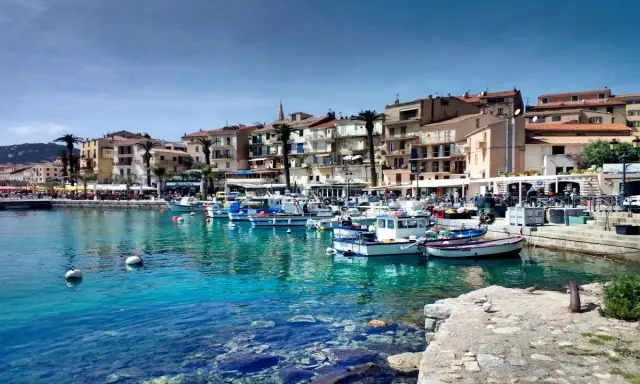 Båtar i en turkos hamn i Calvi, Korsika, med färgglada hus och palmer längs kajen.