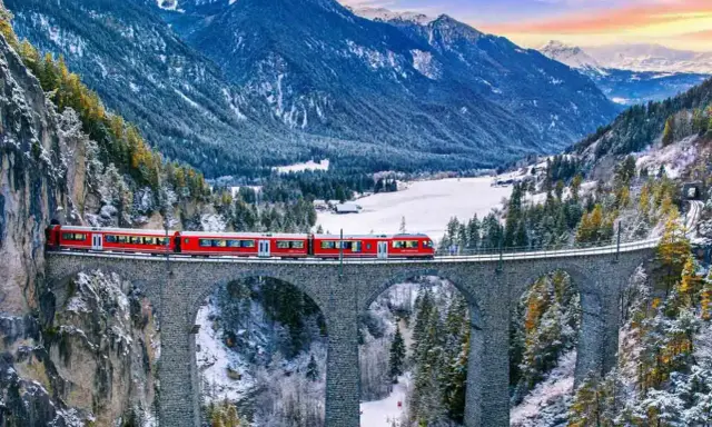 Röda tåget korsar en stenbro över en snöig dal i Schweiz. Fantastiska vyer för din resa till Schweiz tips.