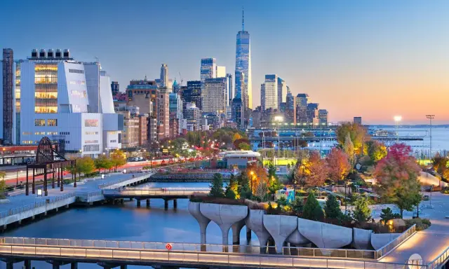 Fakta om New York: En vy över Manhattans skyline vid solnedgången, med den ikoniska One World Trade Center och en modern park vid vattnet.