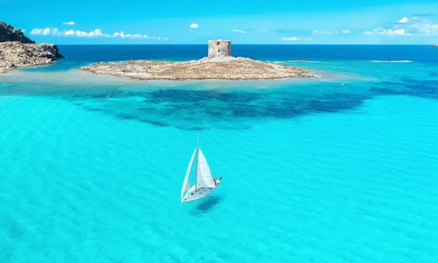 En segelbåt glider fram på turkost vatten mot en gammal vakttorn på en ö. Perfekt för din resa till Sardinien tips!