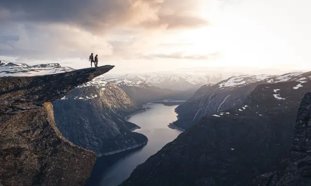 Två personer står på Trolltunga efter en 3 dagars vandring i Norge. Dramatisk utsikt över fjord och snöklädda berg.