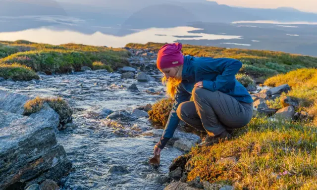 En kvinna fyller sin träkopp vid en bäck under en Abisko vandring dagstur. Solen lyser upp det vidsträckta landskapet.