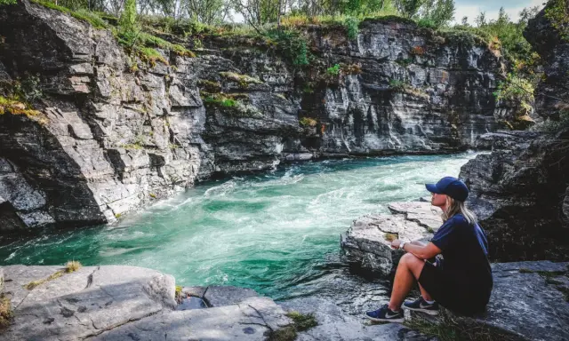 En person sitter vid en forsande turkos flod omgiven av klippor. Ett perfekt utflyktsmål i Mellansverige för naturupplevelser.