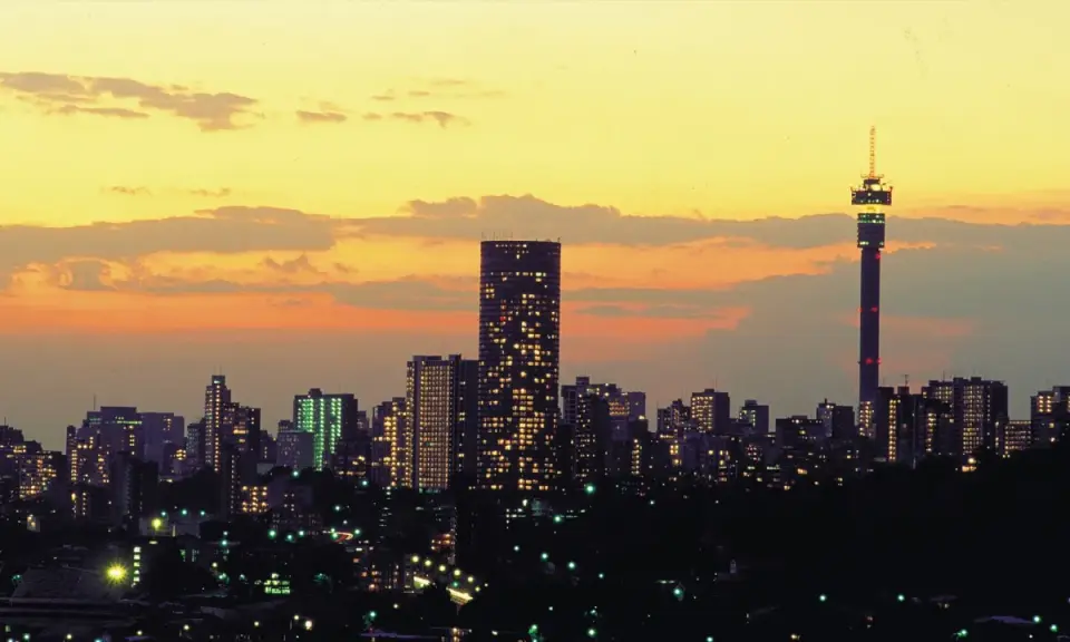 Stadssilhuett i Johannesburg, Sydafrika, med upplysta skyskrapor och ett högt torn mot en gul och orange solnedgångshimmel.