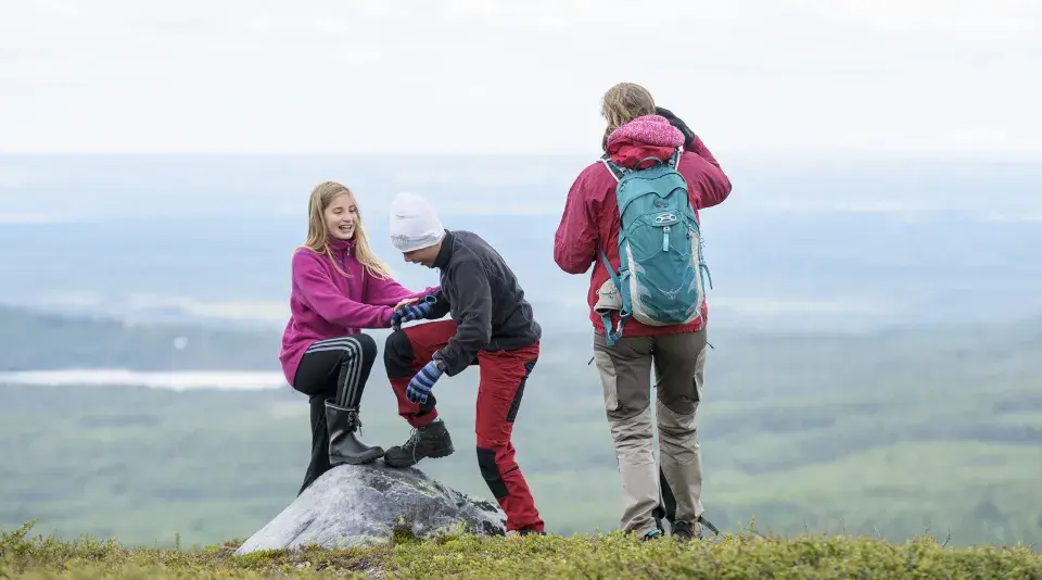 Två barn hjälper varandra upp på en sten under en vandring. En vuxen står bakom med ryggsäcken redo för äventyr.