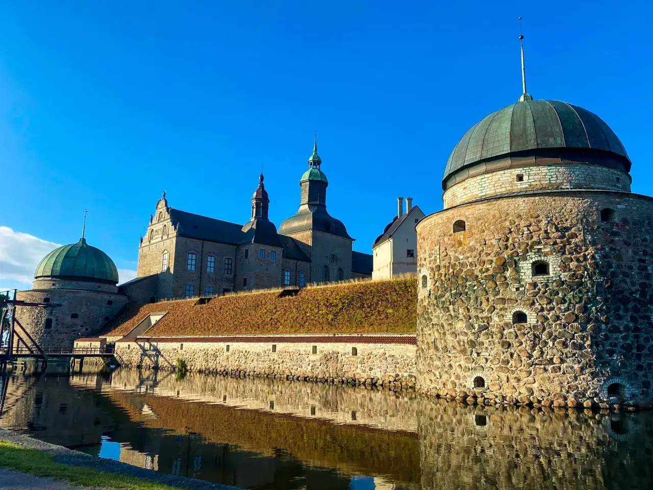 Välkommen till Vadstena! Imponerande slott med kupoler och vallgrav speglar sig i vattnet under en klarblå himmel.