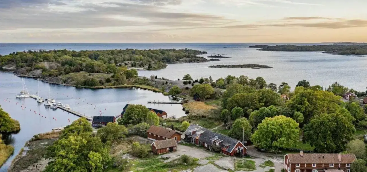 En idyllisk kustvy med röda hus, båtar vid en brygga och grönskande öar. Perfekt för en semester i Blekinge.