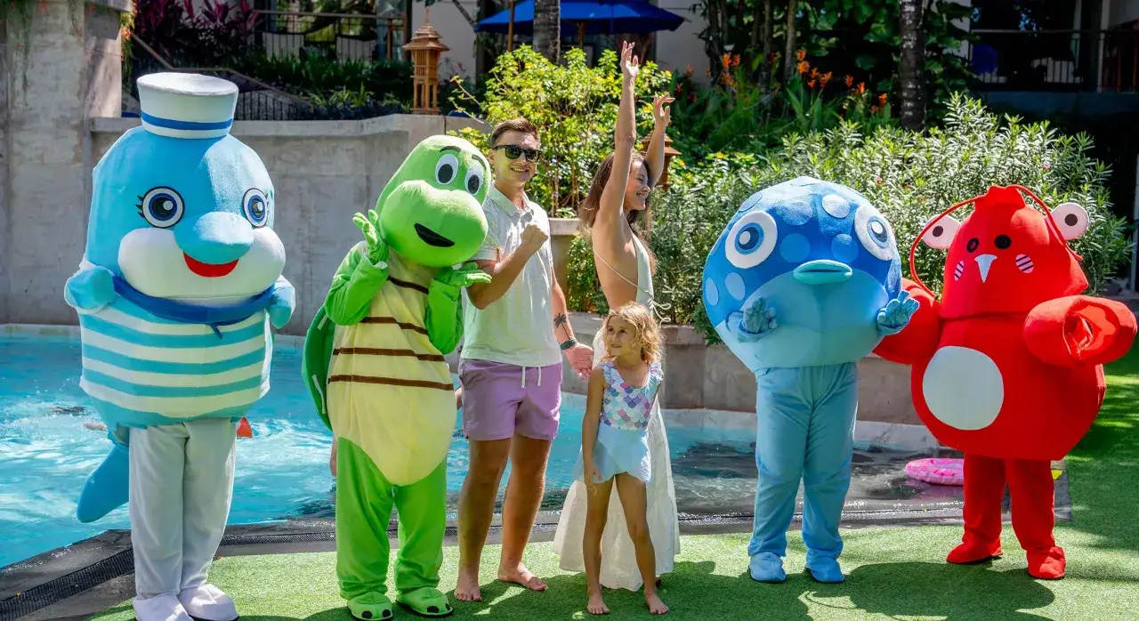 Familjefest i Thailand med barn och maskotar vid poolen. En glad familj njuter av sol och bad med en delfin, sköldpadda, fisk och krabba.