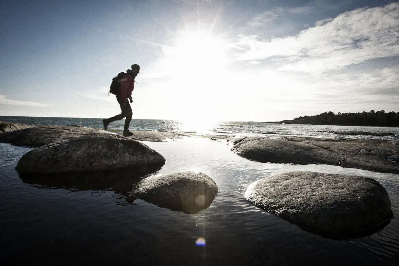 Vandrare med ryggsäck springer över stenar vid havet. En härlig dag för vandring rundslingor Stockholm.