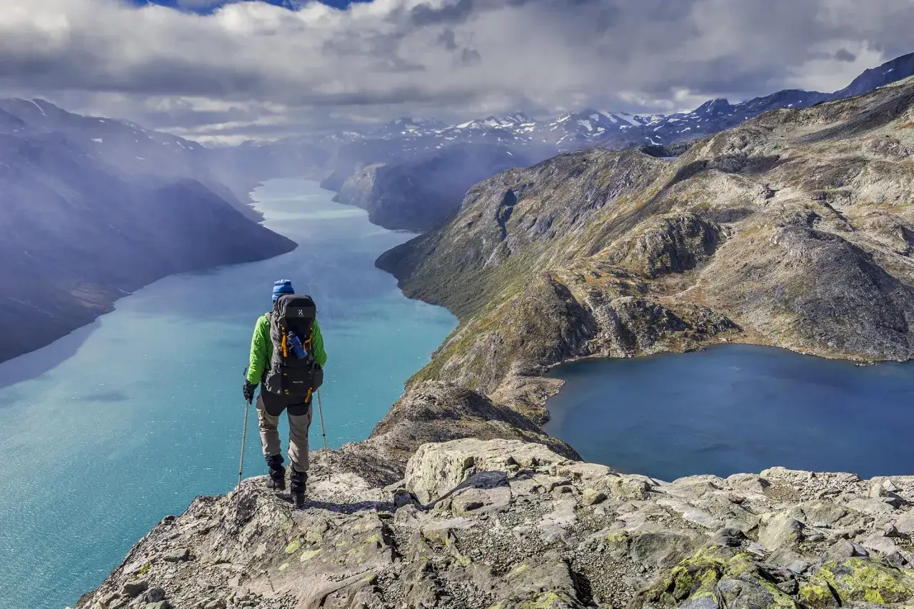 Vandrare med ryggs&auml;ck njuter av en hisnande utsikt &ouml;ver turkosa sj&ouml;ar och sn&ouml;kl&auml;dda berg under en Jotunheimen vandring.