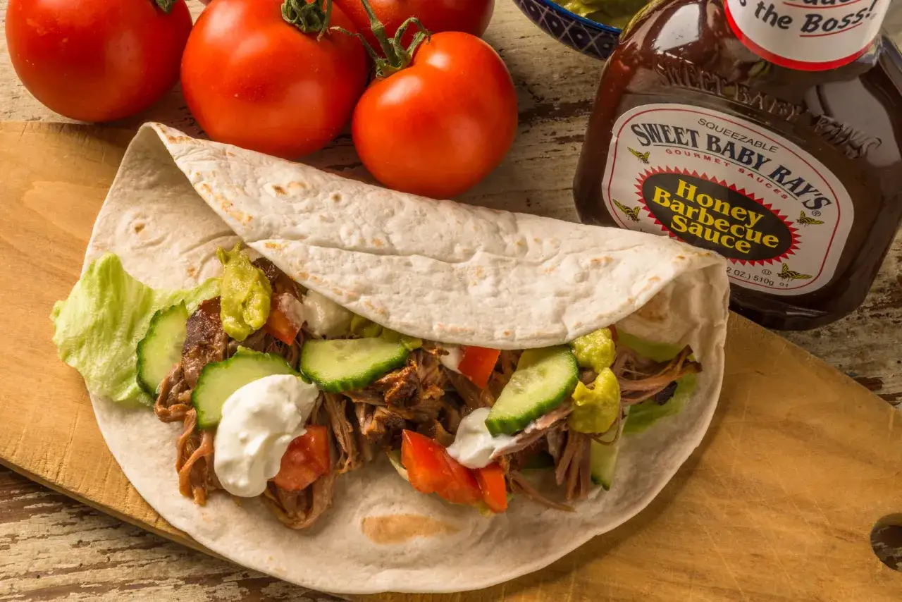 En saftig pulled pork wrap med gurka, tomat och en klick gr&auml;ddfil. Bredvid syns en flaska honungsbarbecues&aring;s.